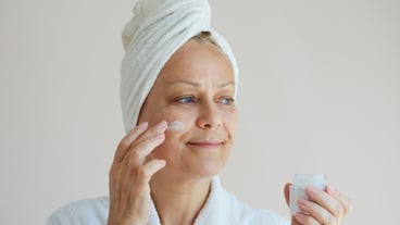 Senior woman in a bathrobe with a white towel on her head uses an antiaging face cream
