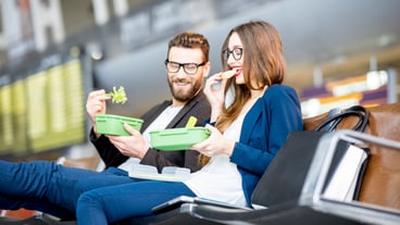 Elegant business couple eating with lunch boxes sitting at the waiting hall in the airport. Having a snack during business trip