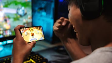 Image of a teenager playing video games with his smartphone and computer in a darkened room, with headphones and a rainbow backlit keyboard.